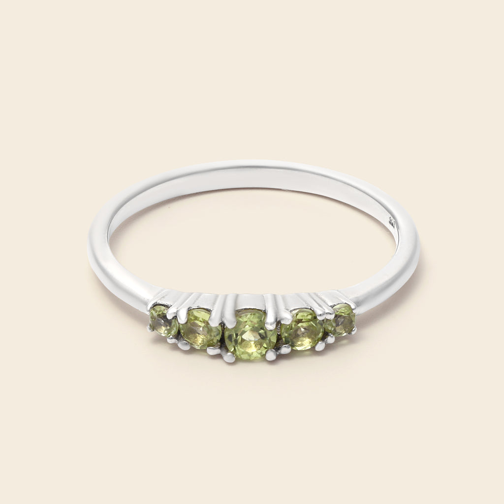 A silver band ring featuring five round-cut light green gemstones in a row, set against a solid light beige background.