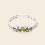 A silver band ring featuring five round-cut light green gemstones in a row, set against a solid light beige background.