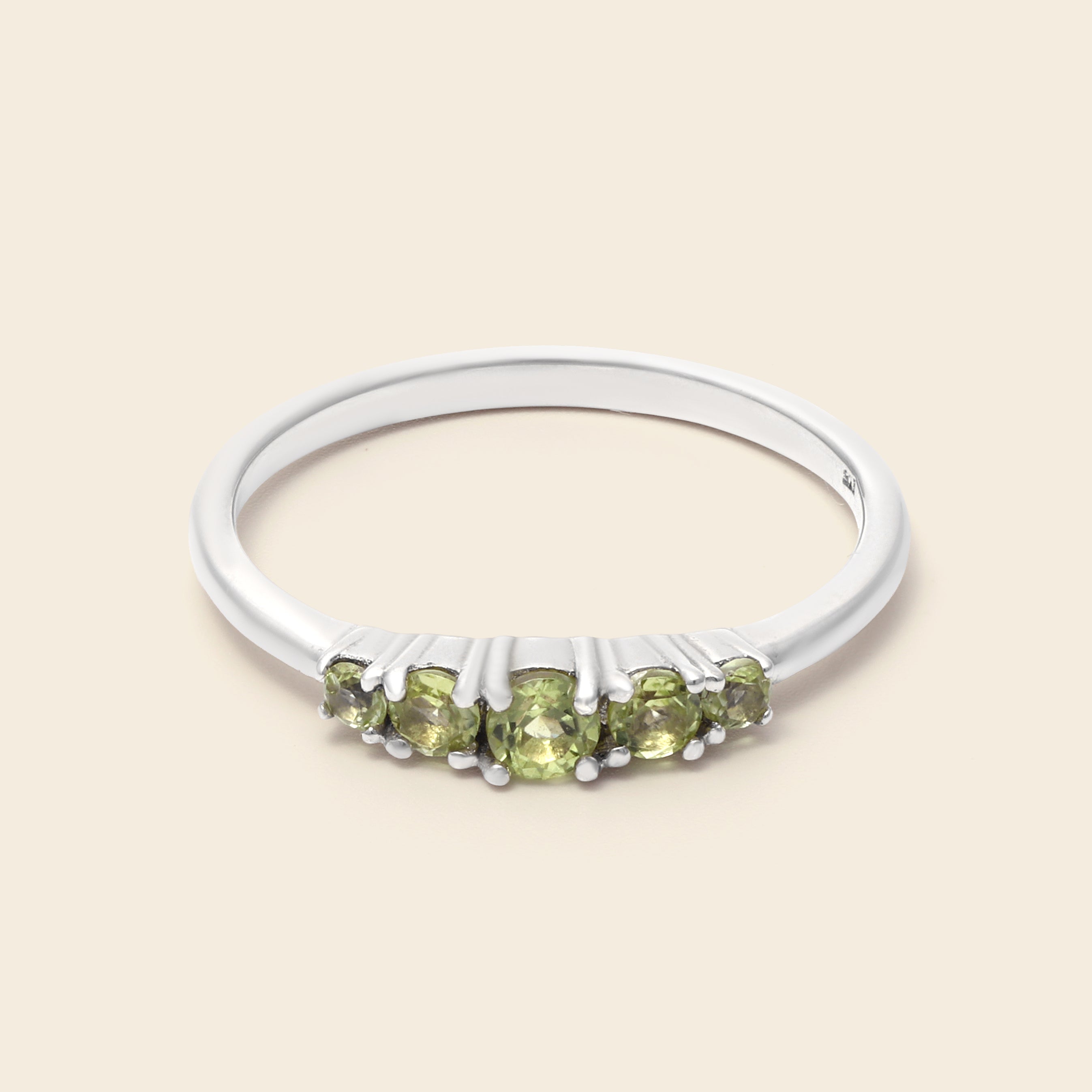 A silver band ring featuring five round-cut light green gemstones in a row, set against a solid light beige background.