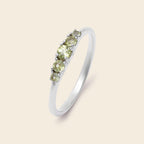 A silver band ring featuring five small round-cut light green gemstones set in a row, shown on a solid pale beige background.