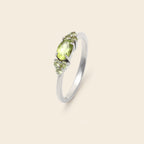 A silver ring featuring a central oval peridot gemstone in a prong setting, flanked by two small round peridot stones, shown on a solid light beige background.