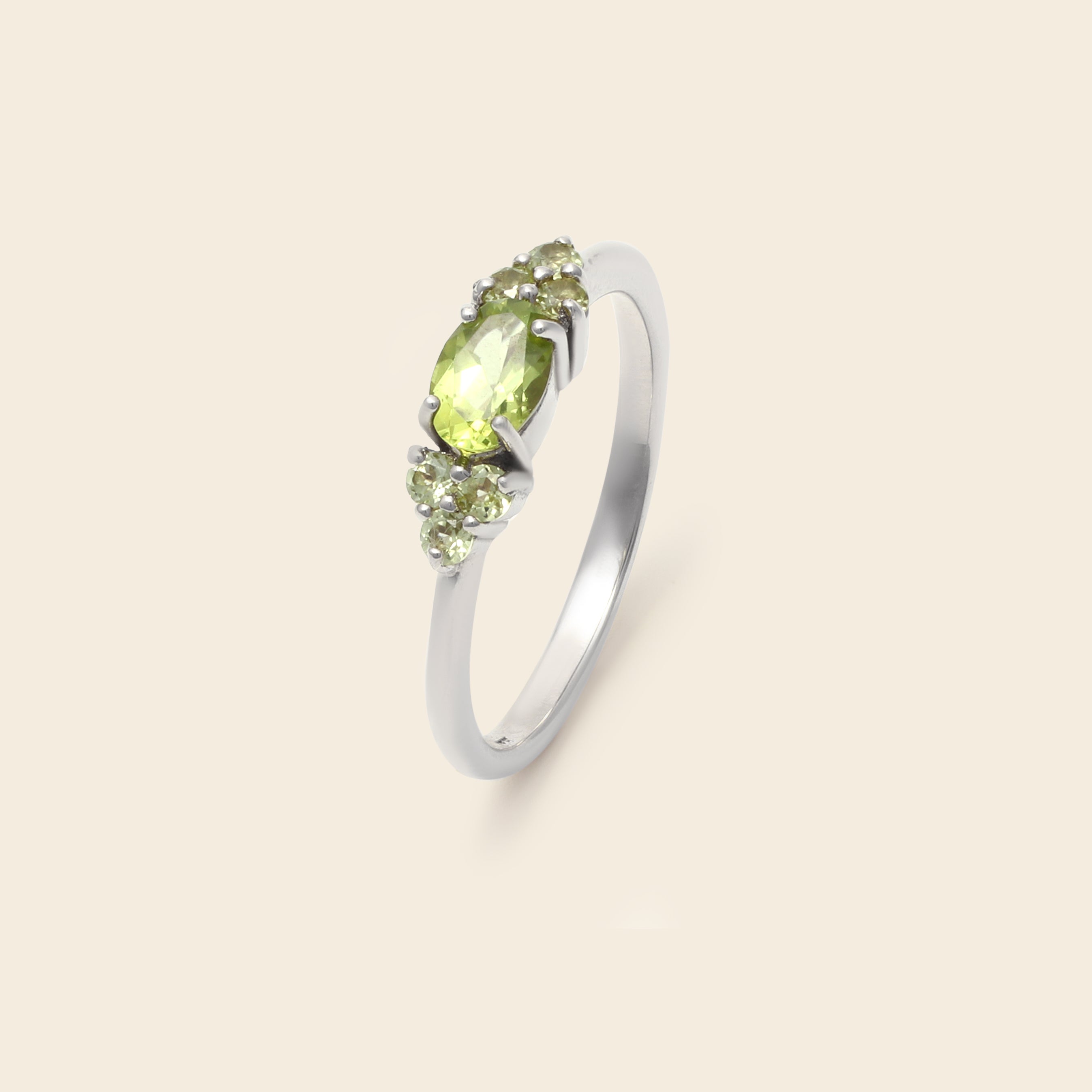 A silver ring featuring a central oval peridot gemstone in a prong setting, flanked by two small round peridot stones, shown on a solid light beige background.