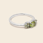A silver three-stone ring featuring oval-cut light green peridot gemstones, shown on a solid light beige background.