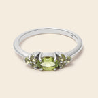 A silver ring featuring a central oval green peridot gemstone flanked by smaller oval-cut green gemstones, set in a polished silver band against a light beige background.