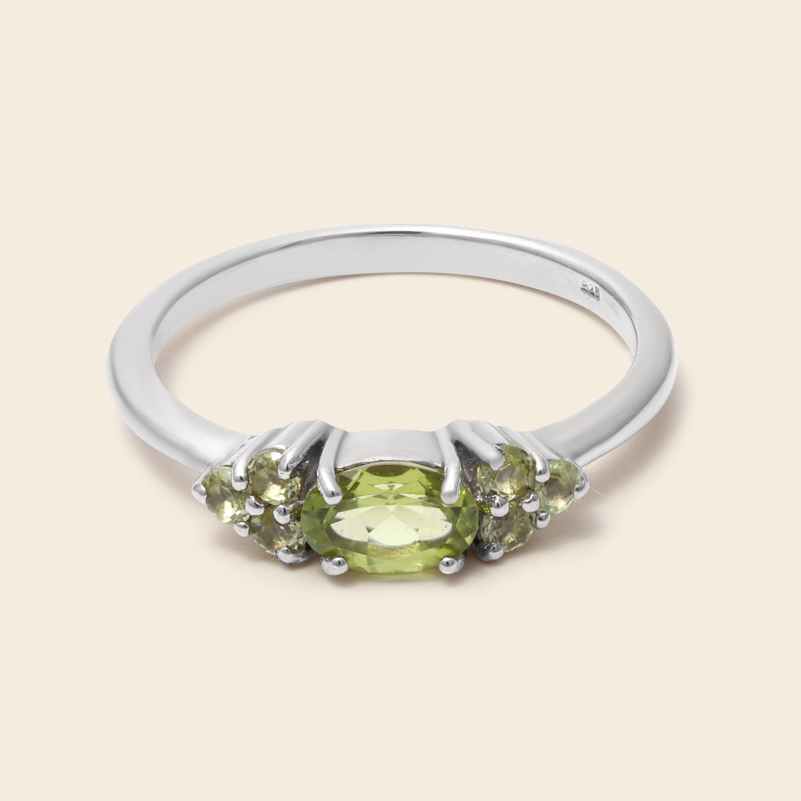 A silver ring featuring a central oval green peridot gemstone flanked by smaller oval-cut green gemstones, set in a polished silver band against a light beige background.
